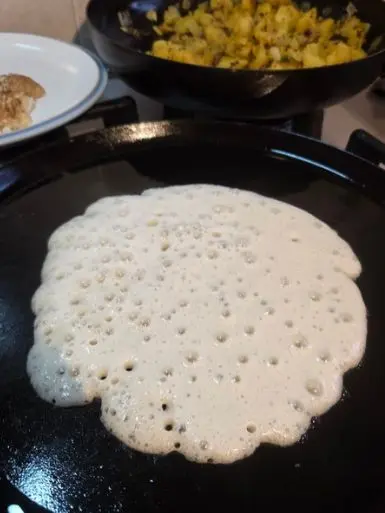 dosa batter bubbling on a frying pan with a pan full of aloo masala in the background